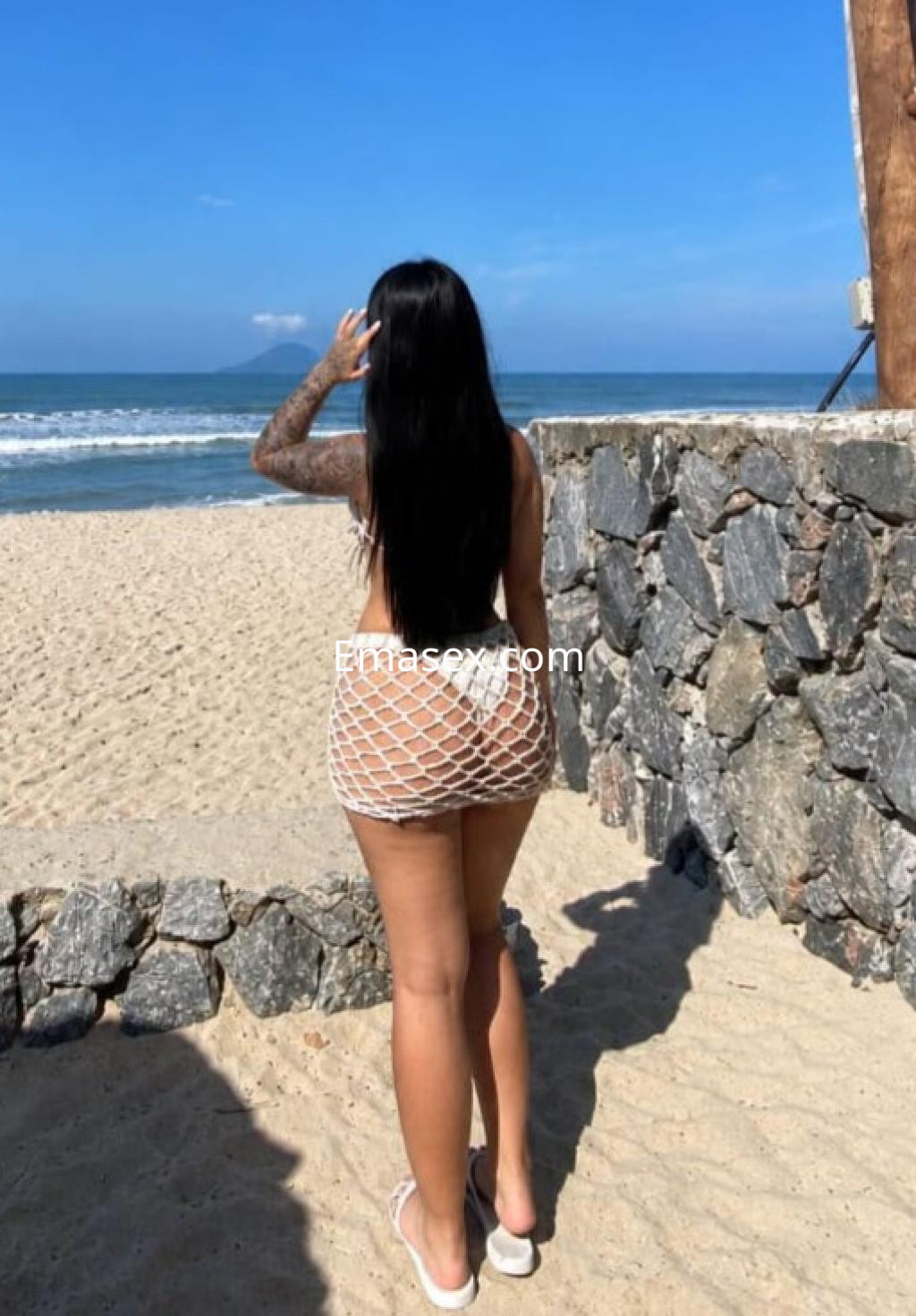 Escort en Coruña, con cabello largo y lacio, luciendo una falda de red blanca, de espaldas frente al mar.