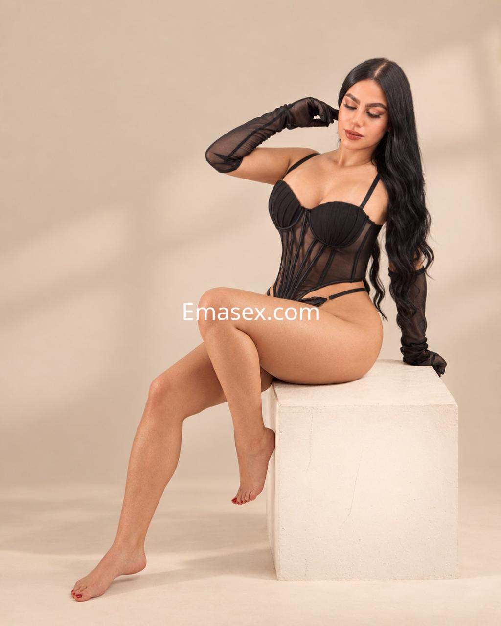 Escort en Bilbao posa sentada sobre un cubo blanco, luciendo un corset negro y guantes largos, con cabello lacio.