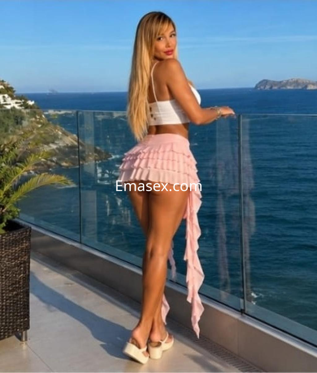 Escort en Irún, de pie junto a un balcón con vista al mar, viste un top blanco y una falda corta rosa.