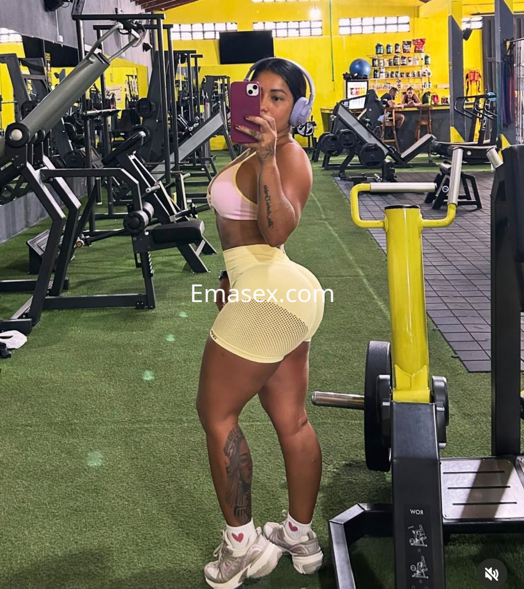 Escort en Alicante, posando en un gimnasio con un top rosa y una falda amarilla, mostrando su figura atlética.