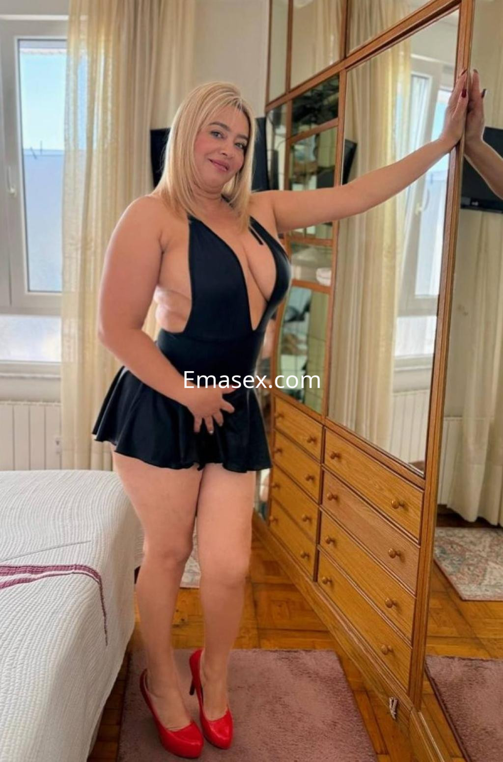 La puta en Donosti lleva un vestido negro de escote profundo y tacones rojos, posando junto a un espejo.