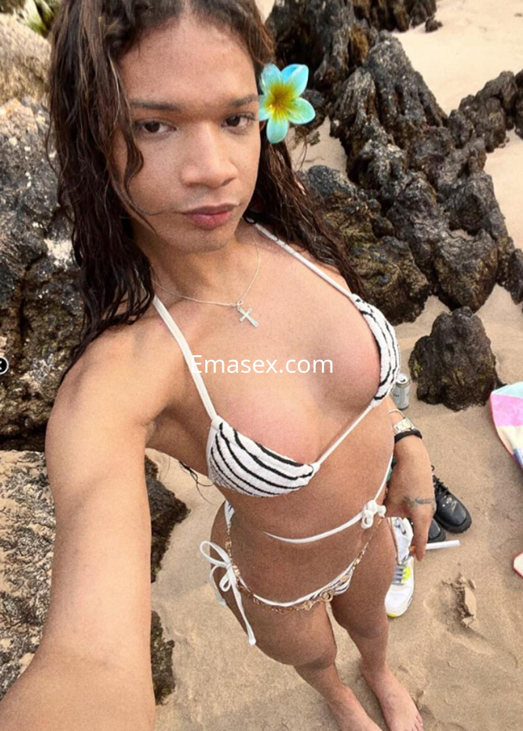 Puta trans en Gijón, tomando una selfie en la playa con bikini de rayas y una flor en el cabello.