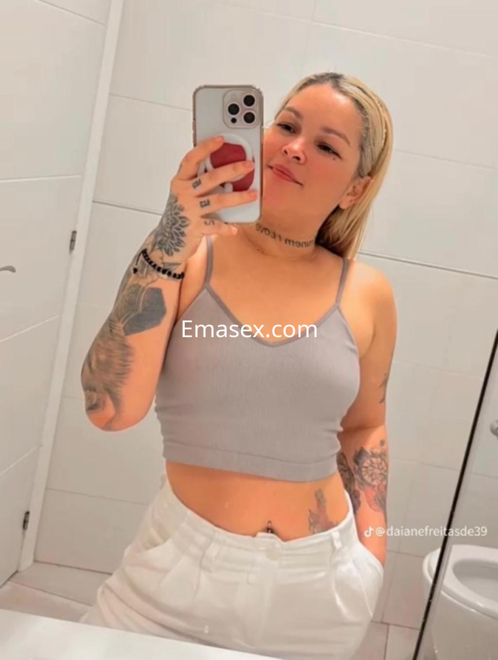 Escort en Lleida con cabello rubio, top gris y pantalones blancos, posando en un baño mientras se toma una selfie.