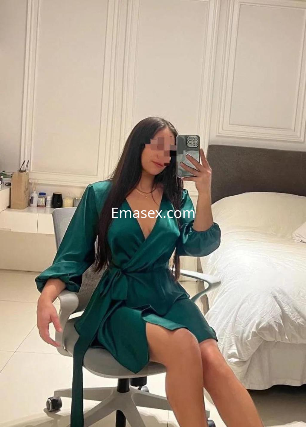 Escort en Lugo posando con un vestido satinado verde, sentada en una silla, frente a una cama y un fondo moderno.