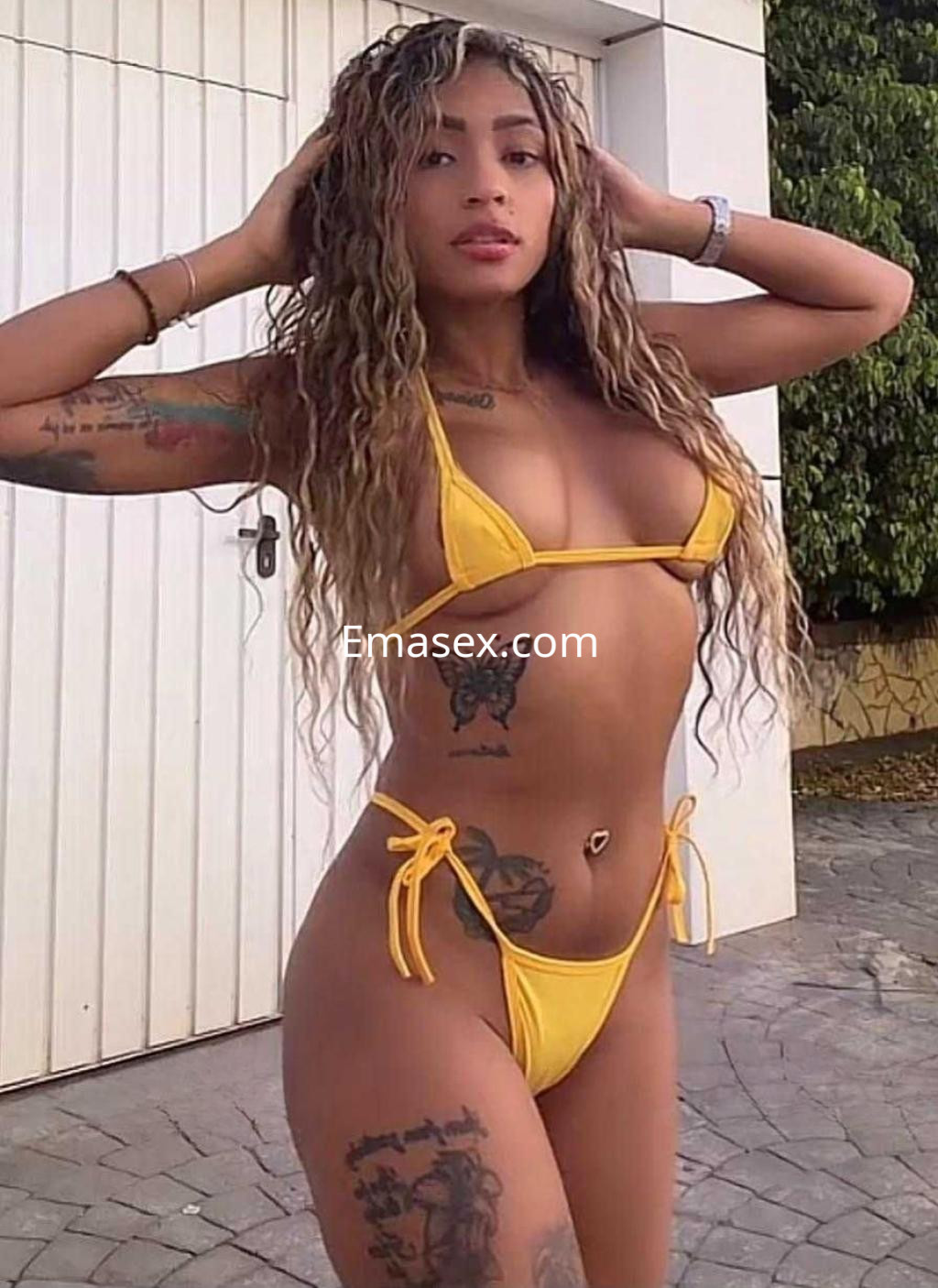 Puta en Málaga, con bikini amarillo, cabello rizado y tatuajes, posando con una mano en la cabeza frente a una pared.