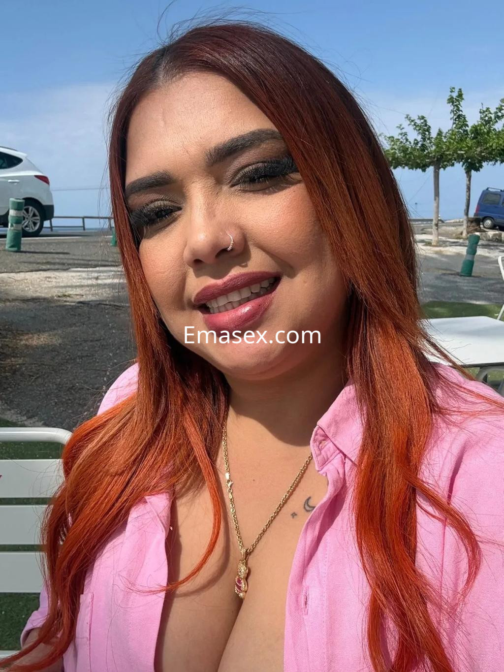 Puta con cabello largo y rojizo, lleva una blusa rosa y sonríe en un entorno al aire libre en Málaga.