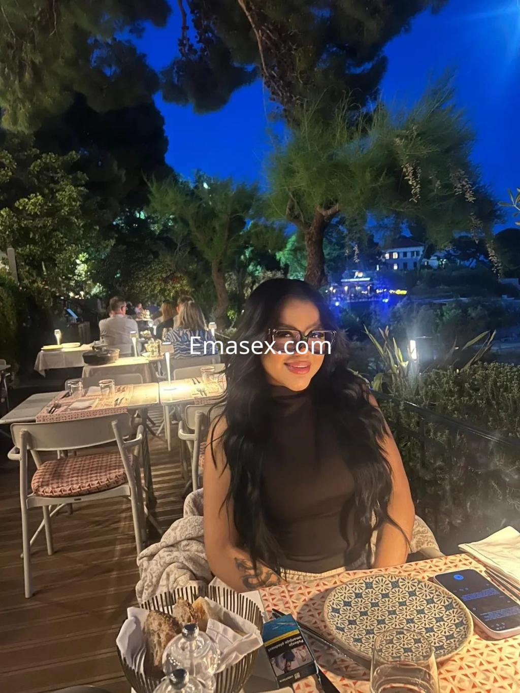 La puta aparece en un restaurante al aire libre en Málaga, luciendo una blusa negra y gafas grandes, sonriendo.