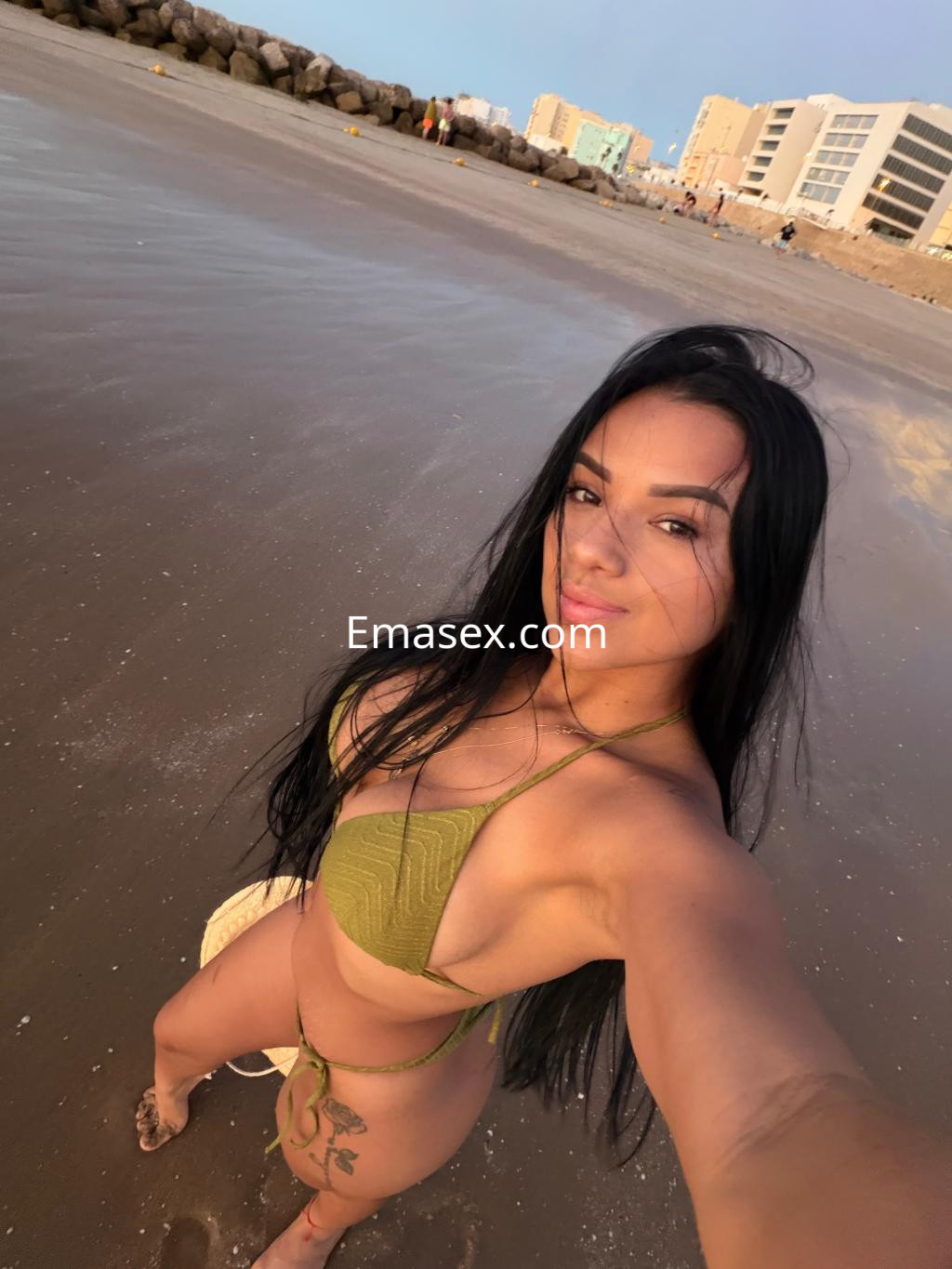Escort en Cádiz, posando en la playa con un bikini verde, cabello largo y suelto, luciendo una sonrisa natural.