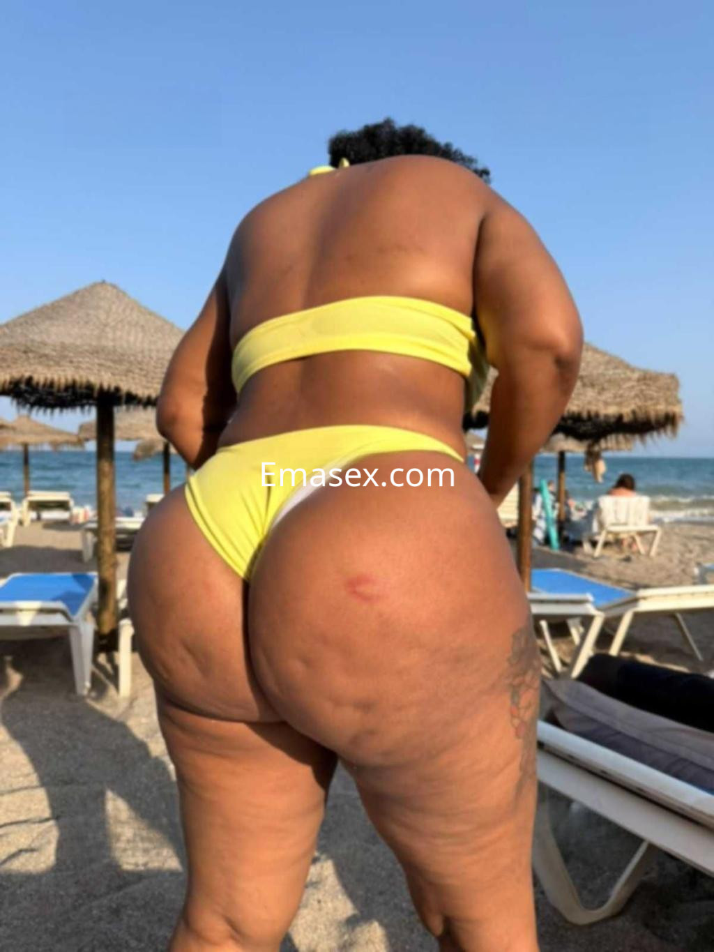 Puta de piel morena con cabello rizado, usando un bikini amarillo en la playa de Ibiza, de espaldas.