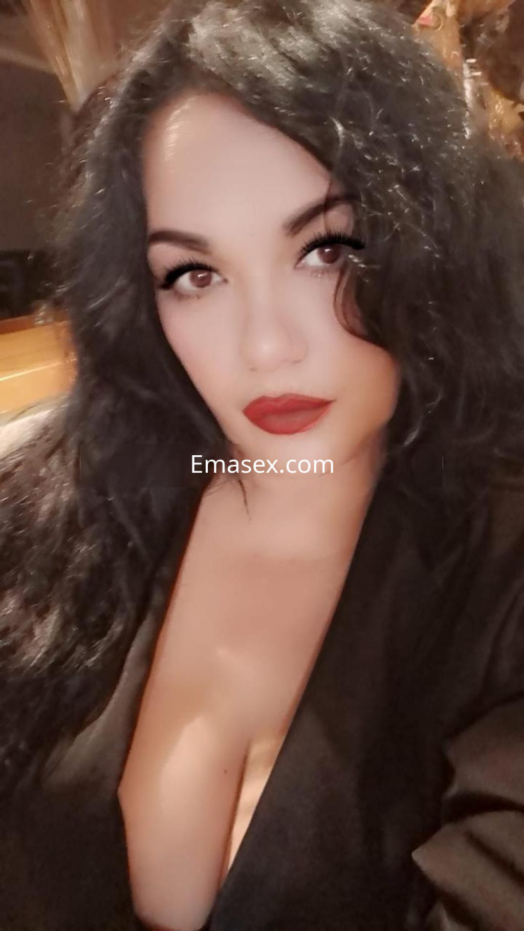 La puta en Lleida luce cabello negro y ondulado, con labios rojos y un escote pronunciado, posando con mirada seductora.