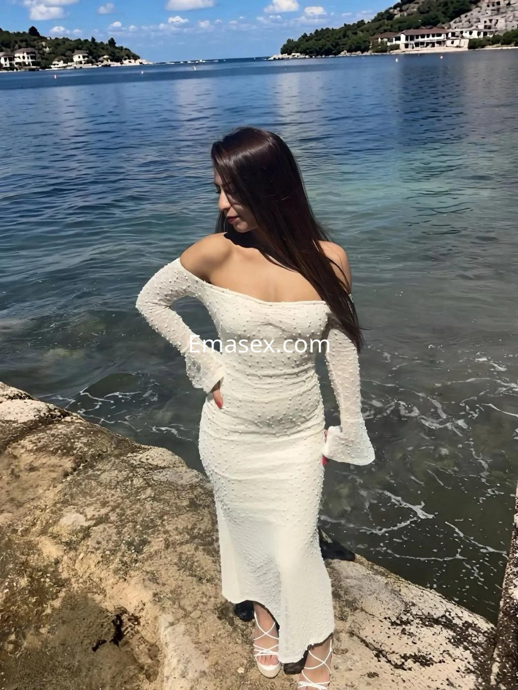 Escort en Málaga con vestido blanco de manga larga, posando junto al mar con una vista de paisaje costero.