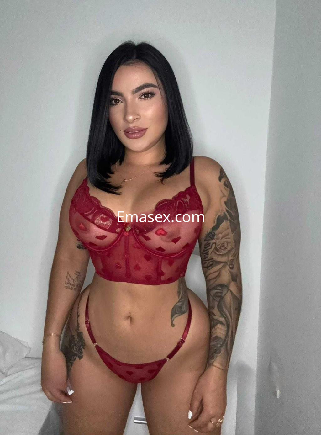 Escort en Cádiz posando con lencería roja transparente, cabello oscuro y tatuajes visibles en los brazos.