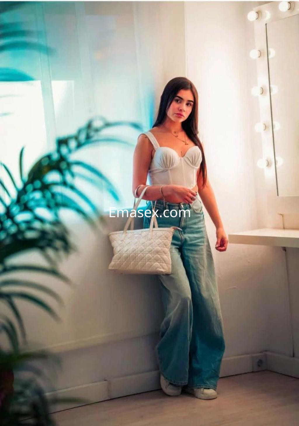 Escort en Alicante con top blanco y jeans anchos, posando junto a un espejo iluminado.