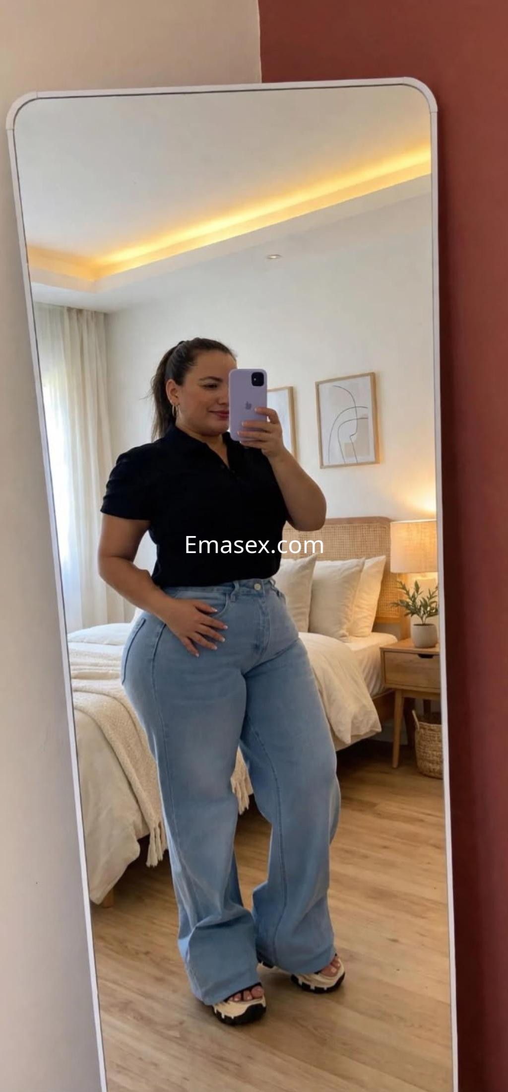 Puta en Burgos posando frente a un espejo, vistiendo una blusa negra y jeans claros, con una habitación de fondo.