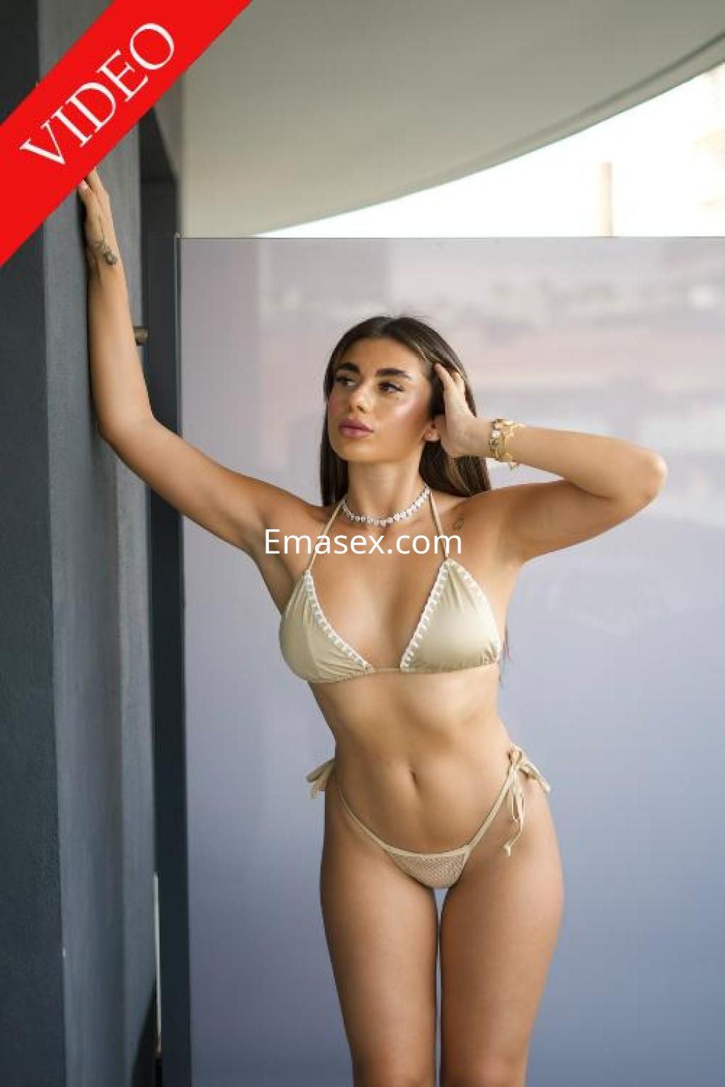 Puta en Ibiza, luciendo un bikini beige mientras se apoya en una pared, con un look veraniego y atractivo.