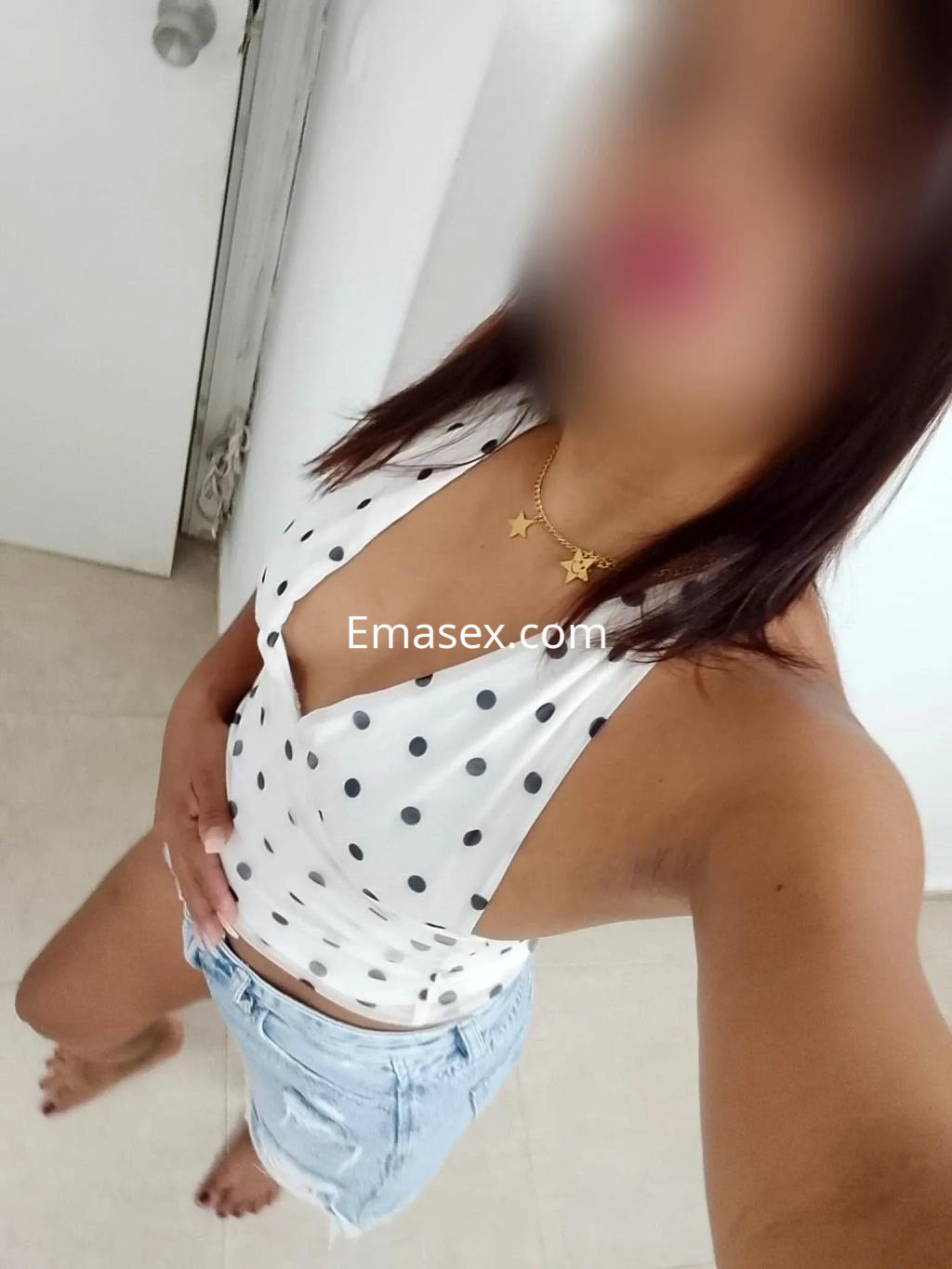 Puta en Cádiz con blusa blanca de lunares y shorts de mezclilla, posando de pie en un ambiente luminoso.