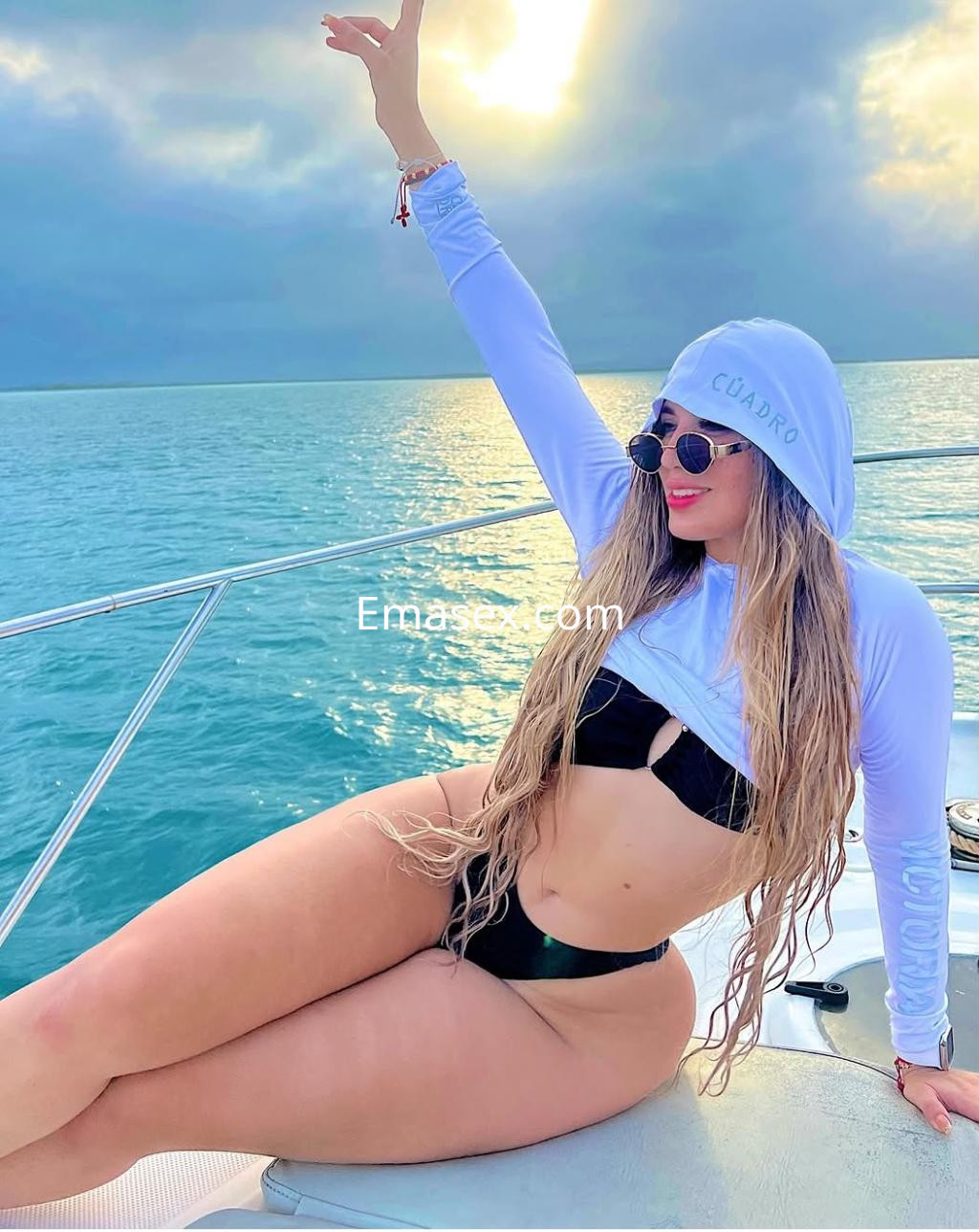 La escort posa en un barco en Granada, vistiendo un top blanco y bikini negro, con el mar y el sol de fondo.