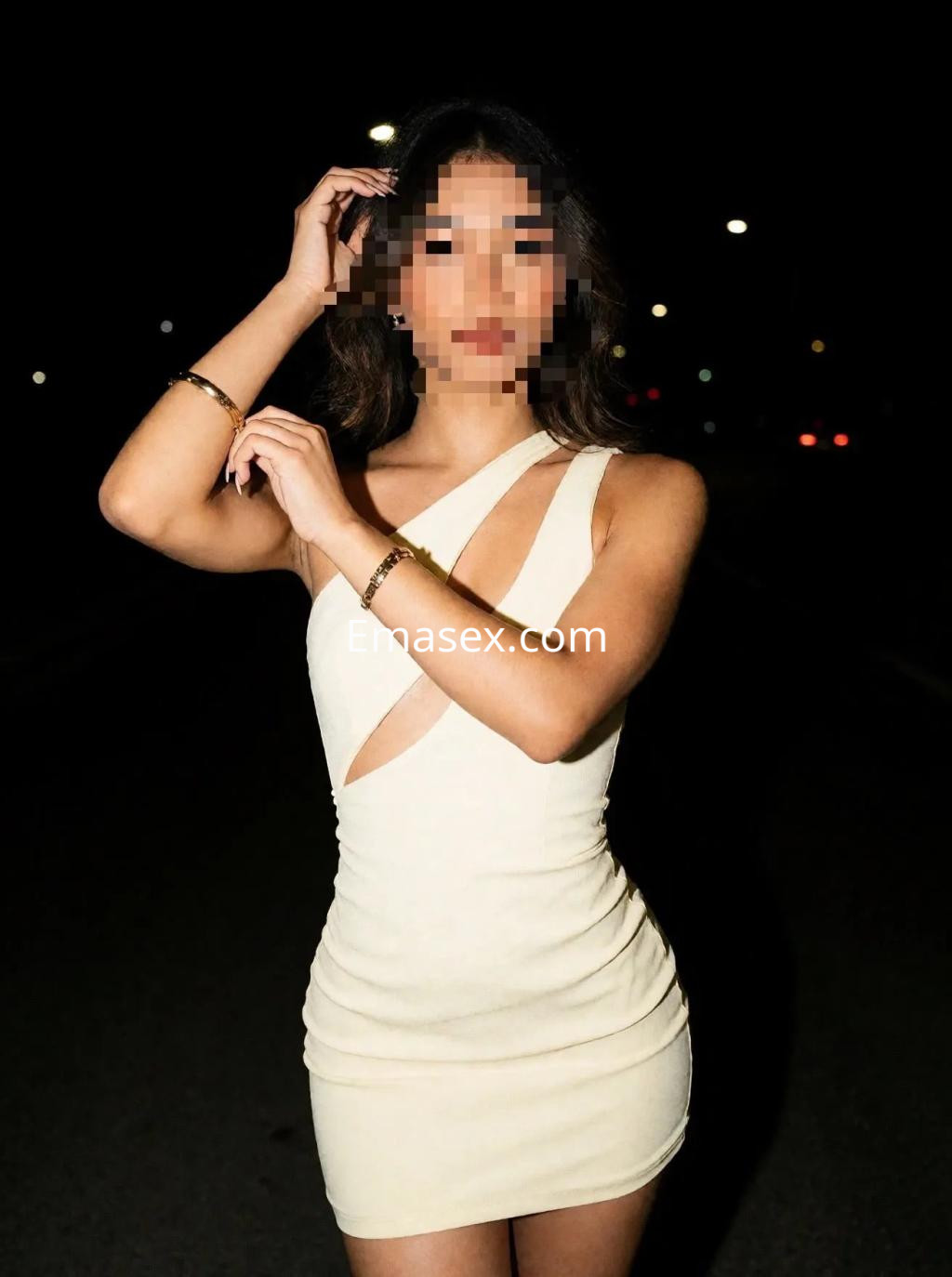 Escort en Madrid, luciendo un vestido corto de color claro con cortes asimétricos, posando al aire libre de noche.