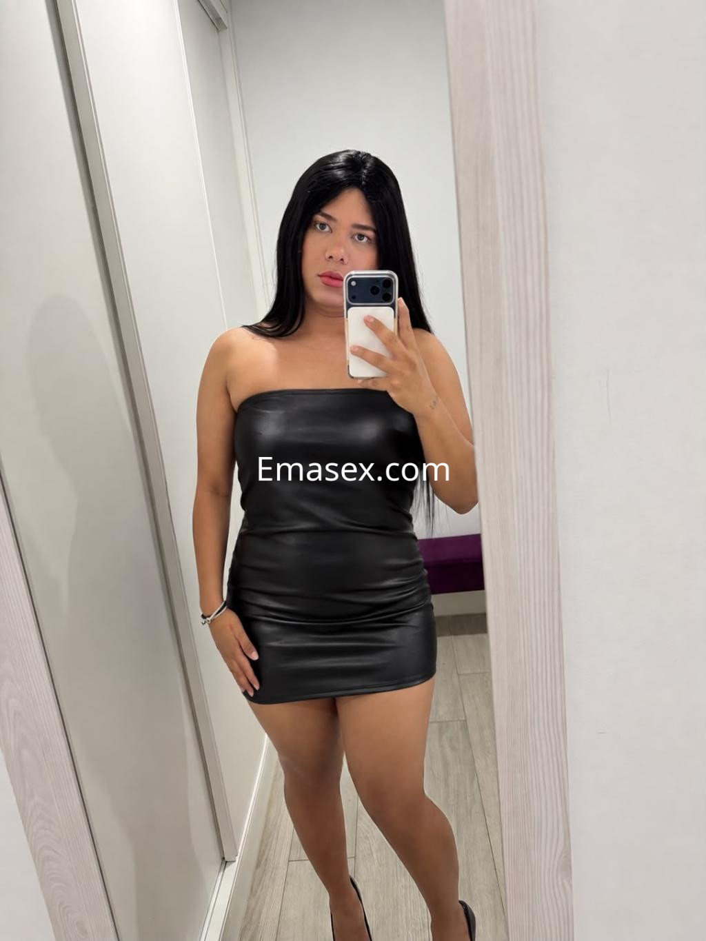Puta trans en Gijón viste un ceñido vestido negro sin mangas, posando frente a un espejo con cabello lacio.