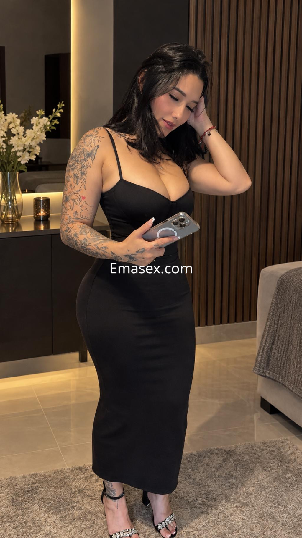 Escort en Irún con vestido negro ajustado, cabello oscuro y tatuajes visibles, posando con un teléfono en la mano.