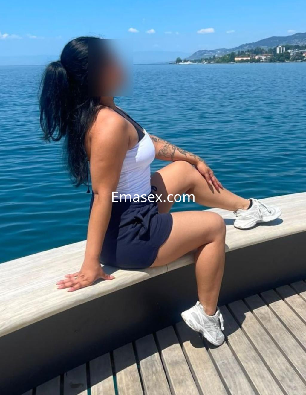 Escort en Madrid con una blusa blanca y shorts oscuros, posando en un muelle frente al mar.
