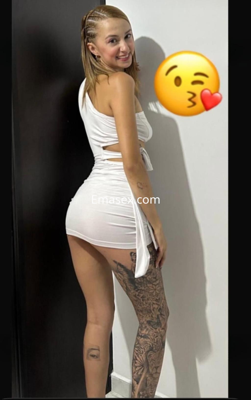 Escort en Murcia posando de lado, con cabello corto y rubio, vestida con un vestido blanco ajustado y tatuajes en la pierna.