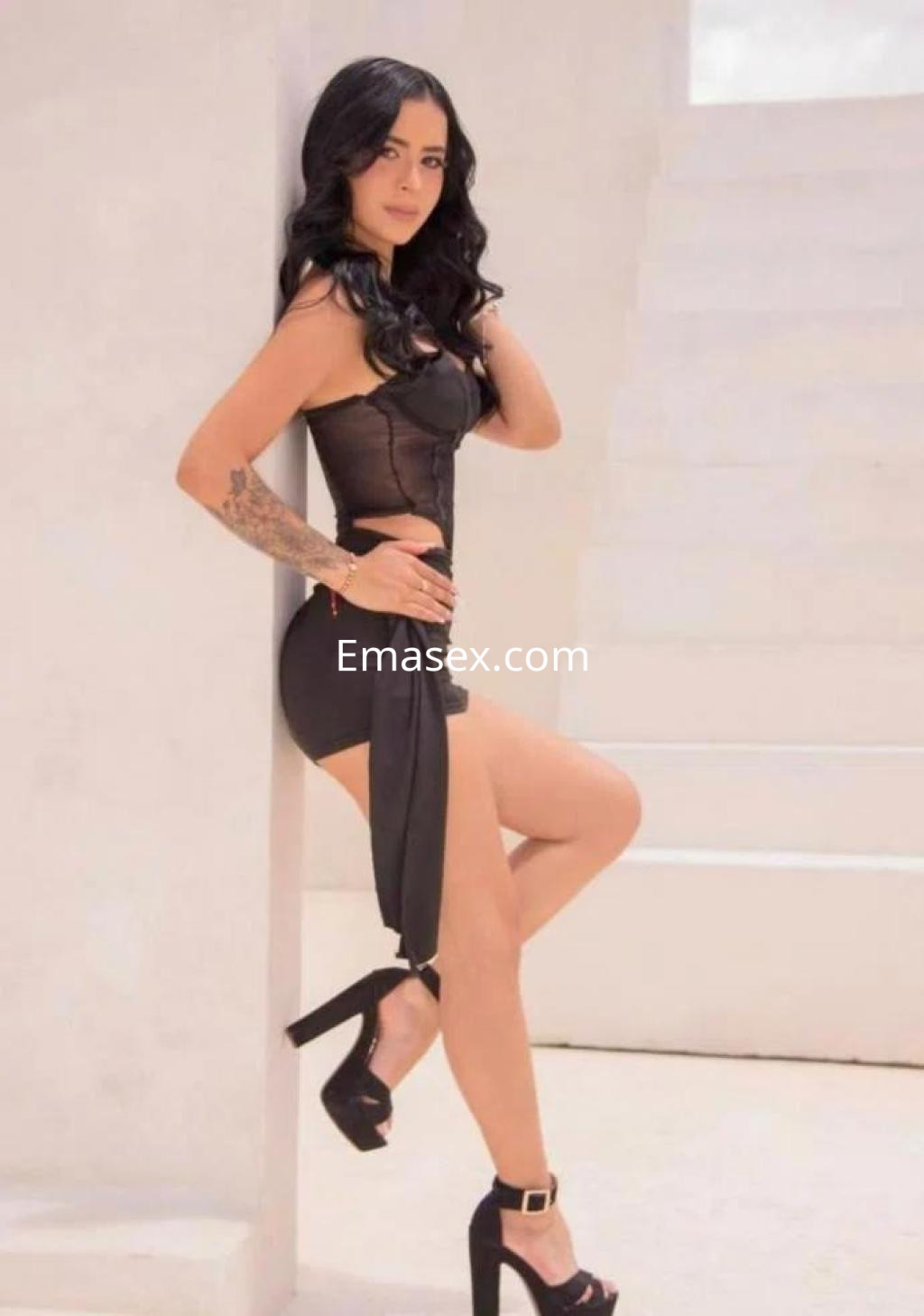 Escort de Palma de Mallorca posando de pie con un vestido negro ajustado y tacones altos en un entorno iluminado.