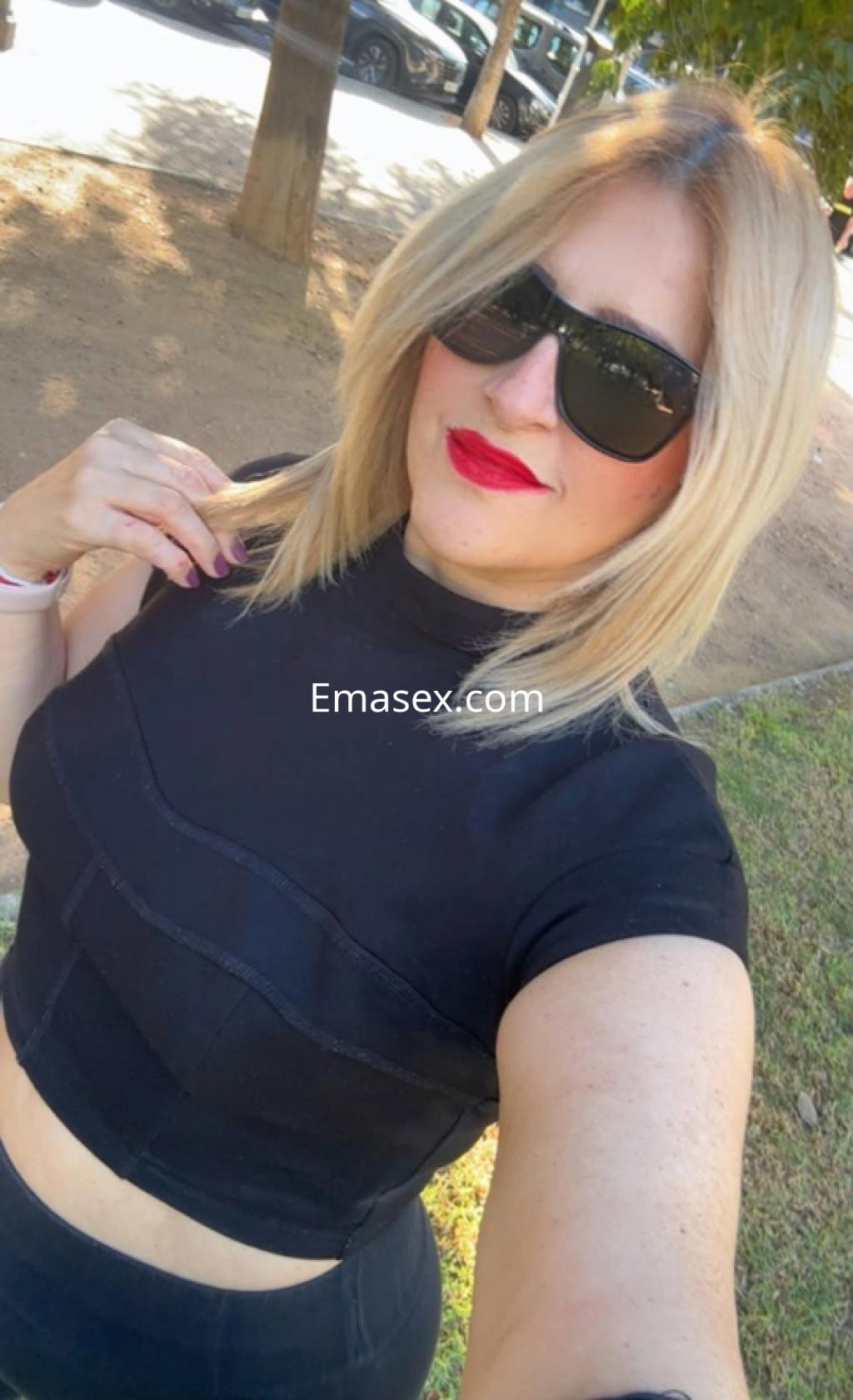 Escort en Córdoba con cabello rubio, usando una blusa negra y gafas de sol, posando en un entorno al aire libre.