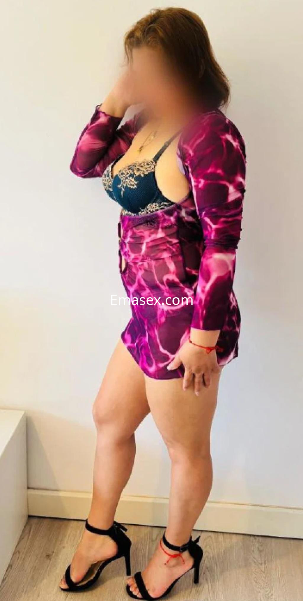 Puta de pie, luciendo un vestido corto con estampado en tonos morados y sandalias de tacón, con cabello castaño.