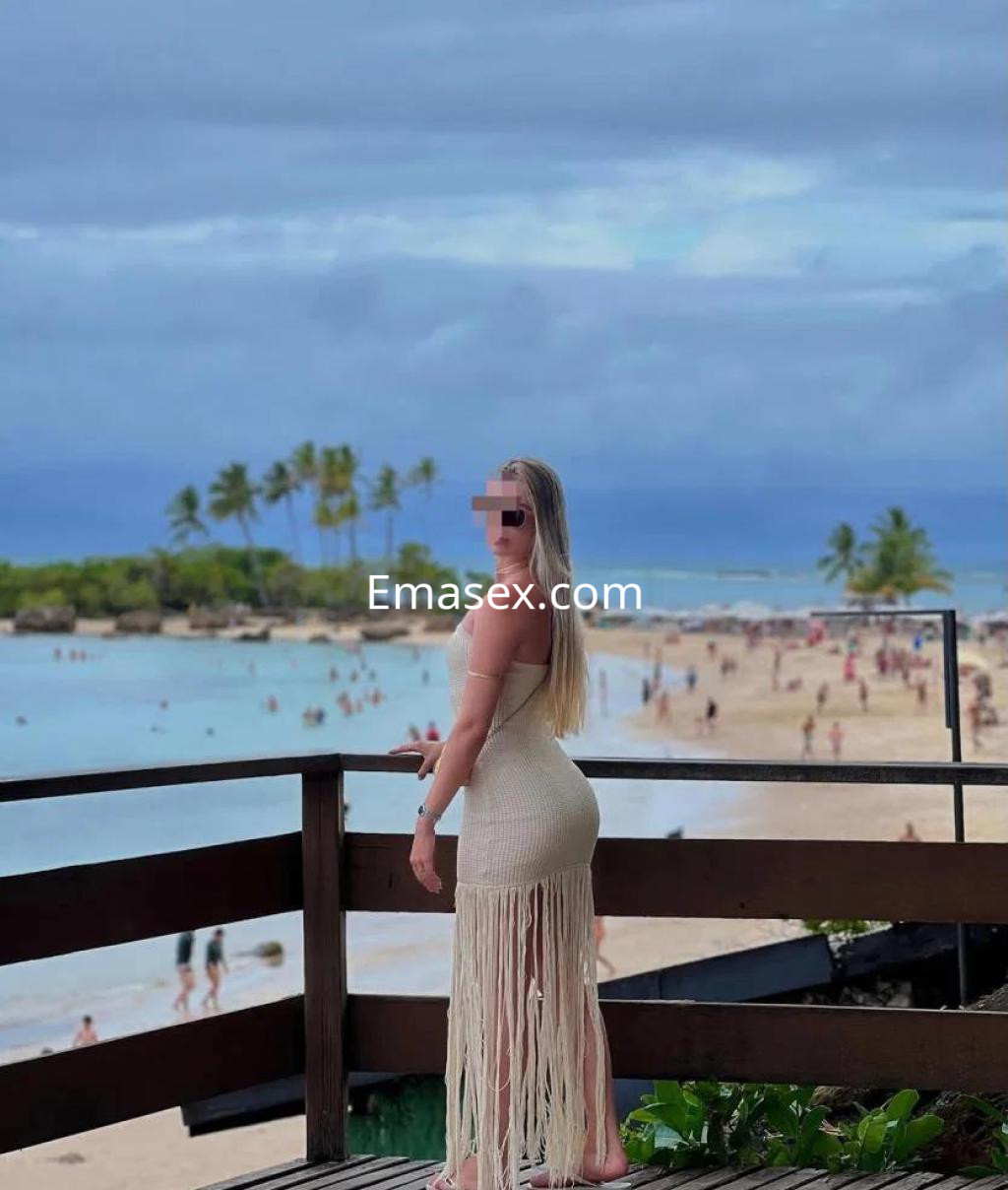 La puta está de pie en un mirador con vista a la playa en Tenerife, luciendo un vestido de tirantes y flecos.