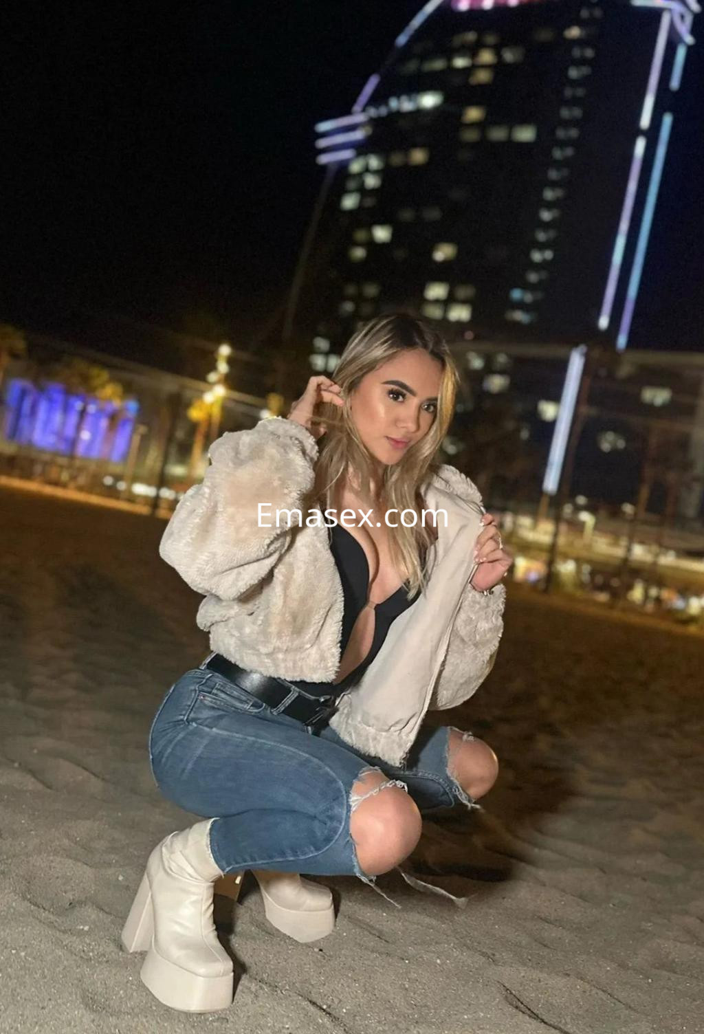 Puta en Palma de Mallorca, con cabello largo y rubio, viste un top negro y jeans rasgados, posando en la playa.