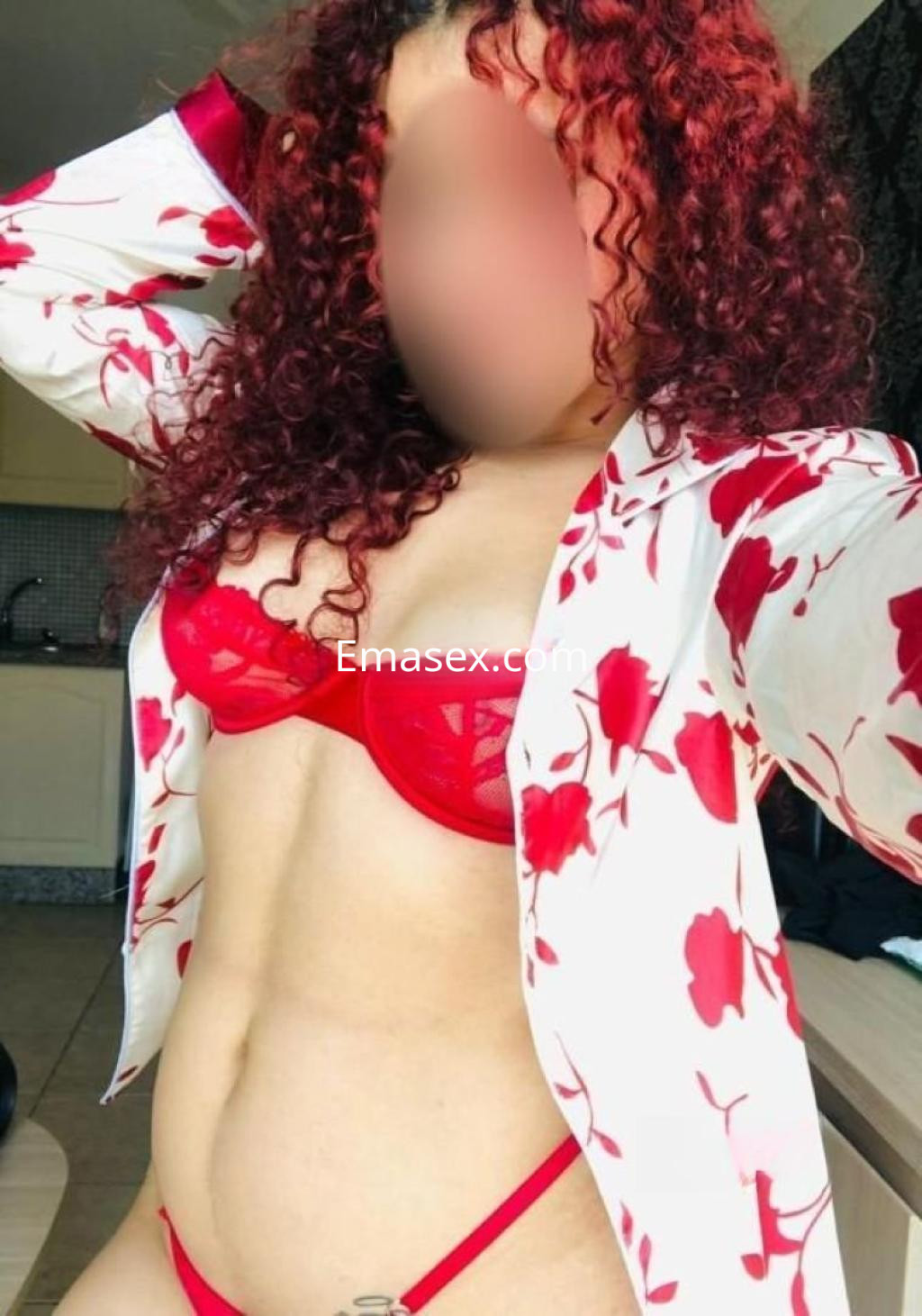 Escort con cabello rizado y rojo, vistiendo un sujetador y tanga rojos, posando con una camisa blanca con flores rojas en Santander.