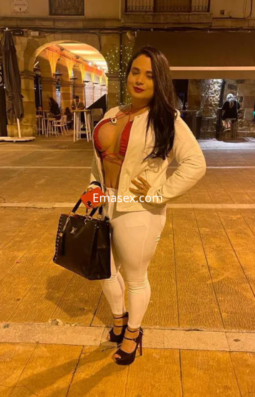 Escort trans en Santander con top rojo y leggings blancos, sosteniendo un bolso negro en una calle iluminada.