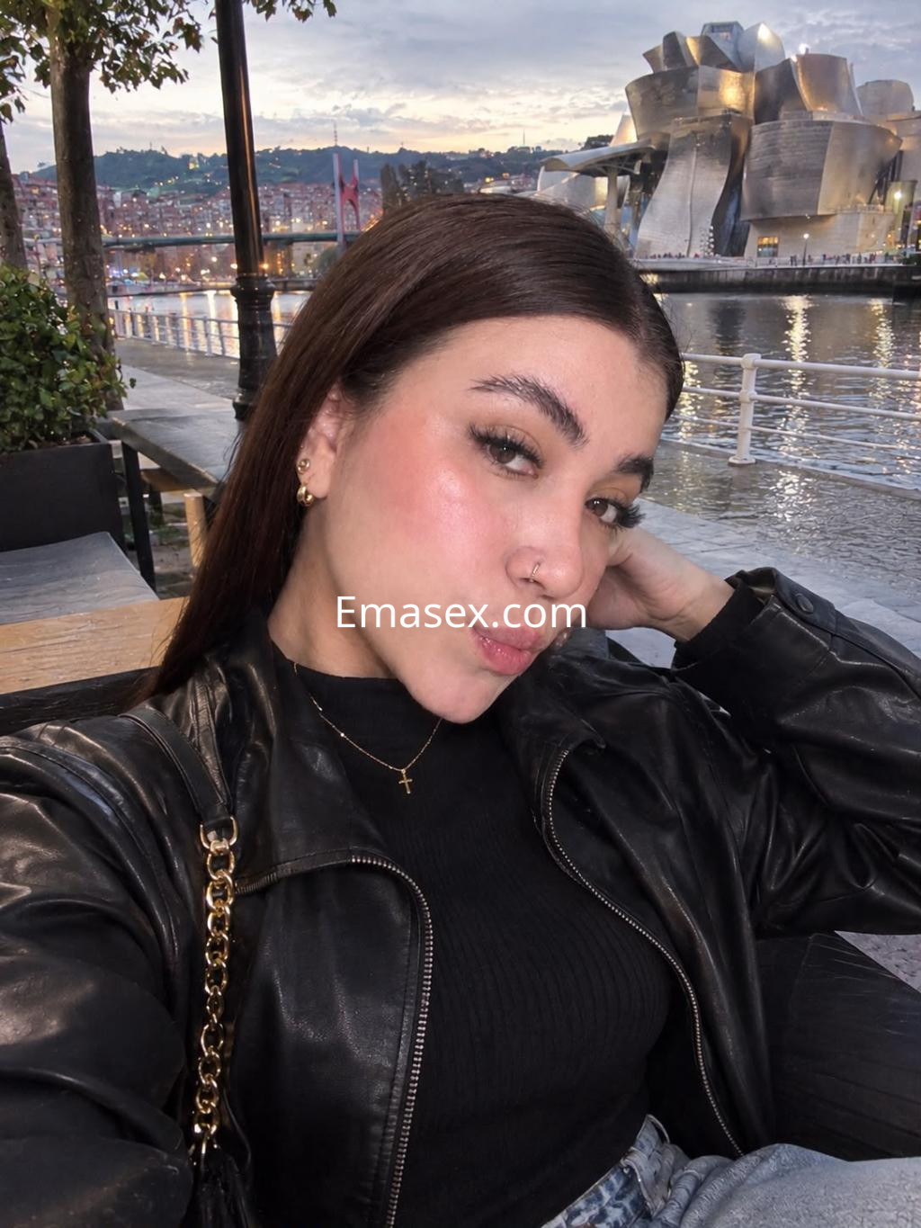 Escort en Bilbao posa al aire libre con una chaqueta de cuero negra y un top negro, frente al río y el museo.