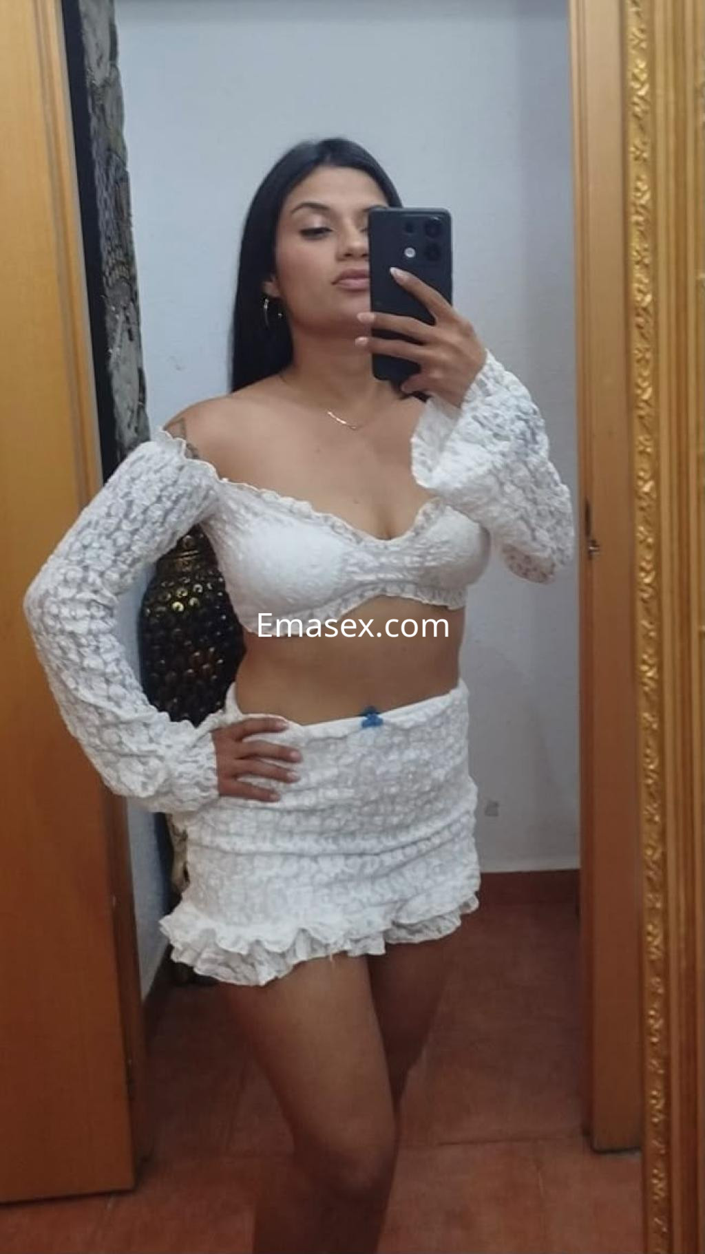 Escort en Valencia posa frente al espejo con un conjunto blanco de top y falda corta, mostrando su figura.