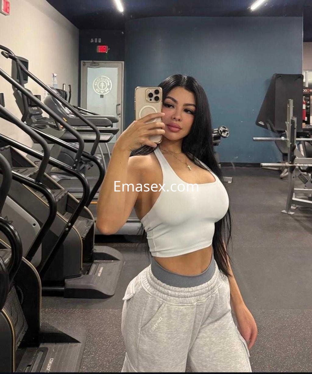 Escort en Valladolid en un gimnasio, vestida con top blanco y pantalones deportivos grises, tomando un selfie.