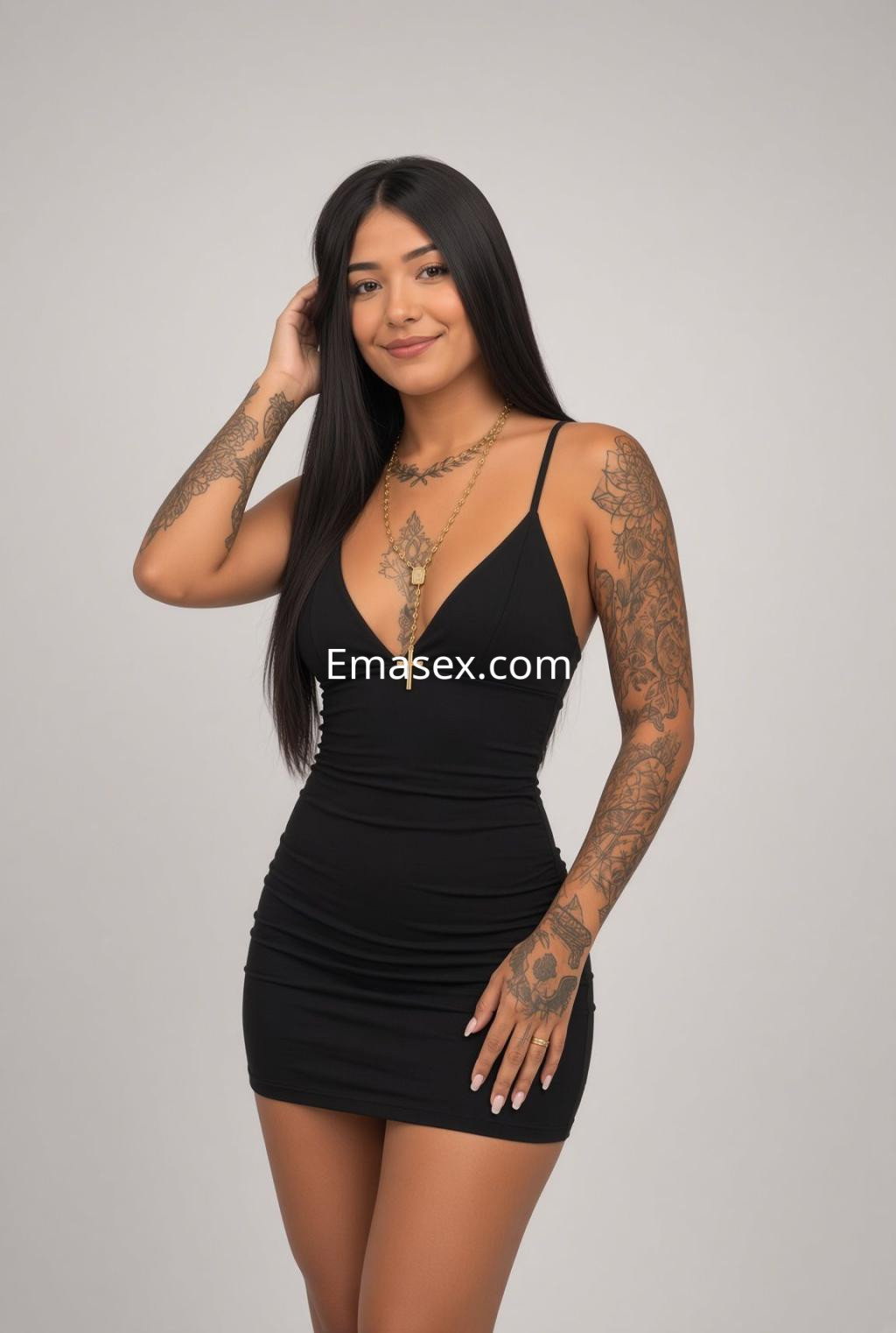 Escort en Sevilla con vestido negro ajustado y tatuajes visibles, posando con una mano en su cabello.