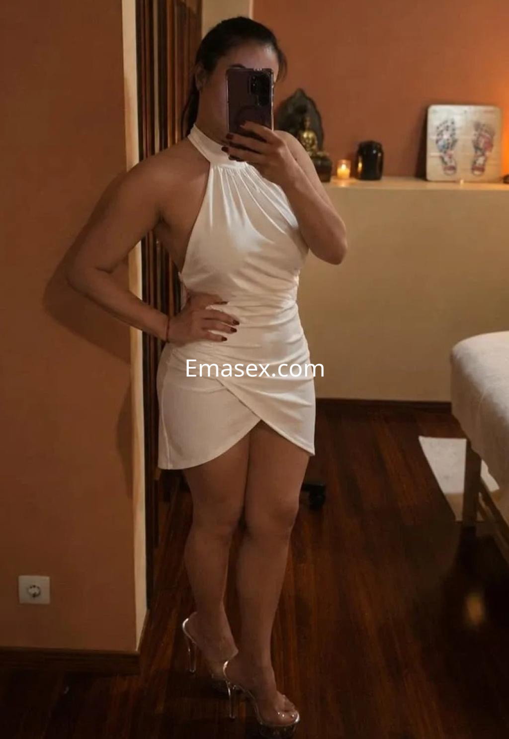 Escort en Vigo con un vestido blanco sin mangas y tacones transparentes, posando frente al espejo en un ambiente cálido.