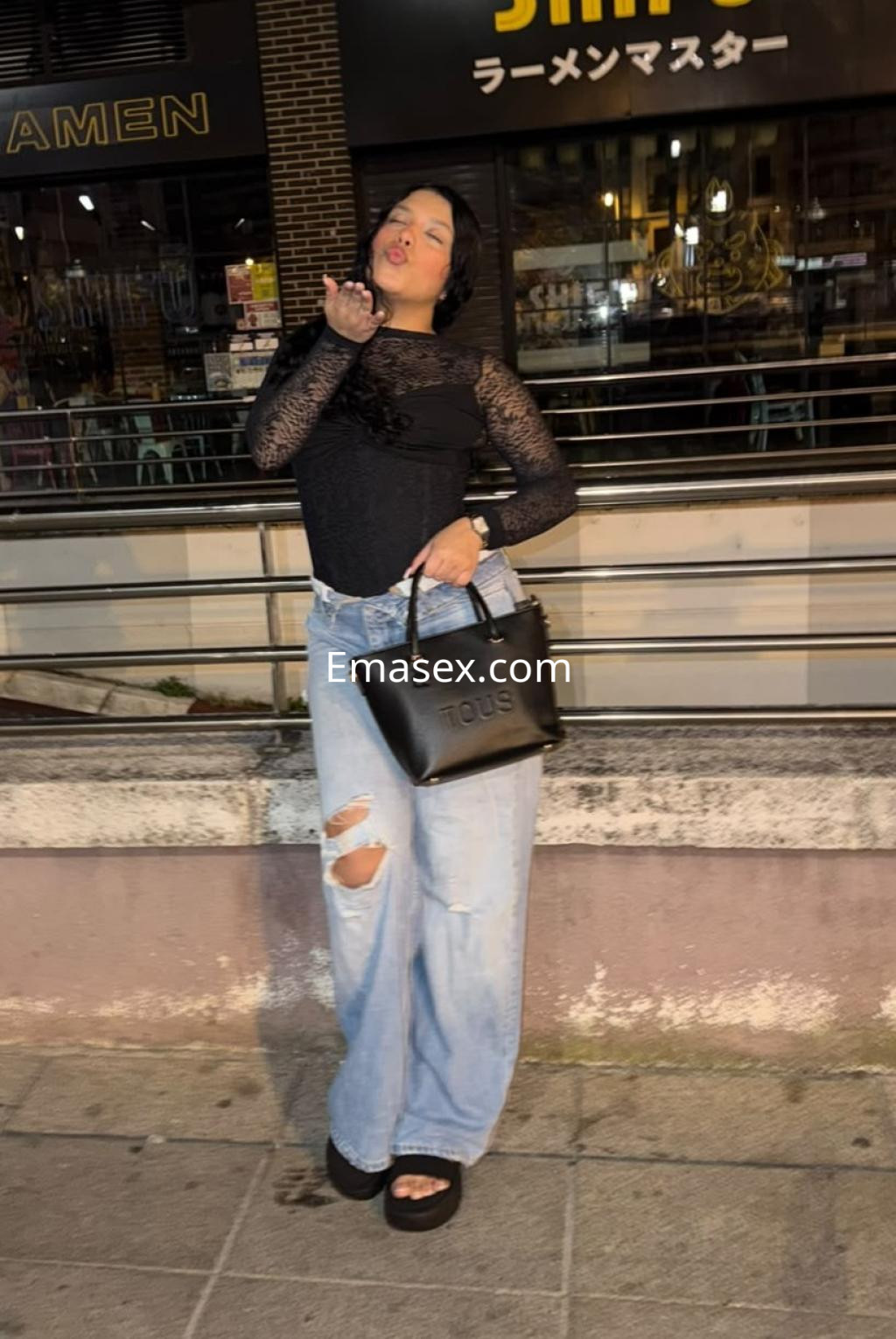 Escort en Logroño, con cabello lacio y oscuro, viste una blusa negra y jeans holgados, posando con un bolso.