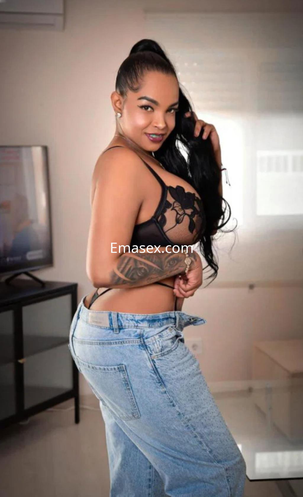 Escort en Zaragoza con cabello largo y oscuro, vistiendo un sujetador negro y jeans ajustados, posando con confianza.