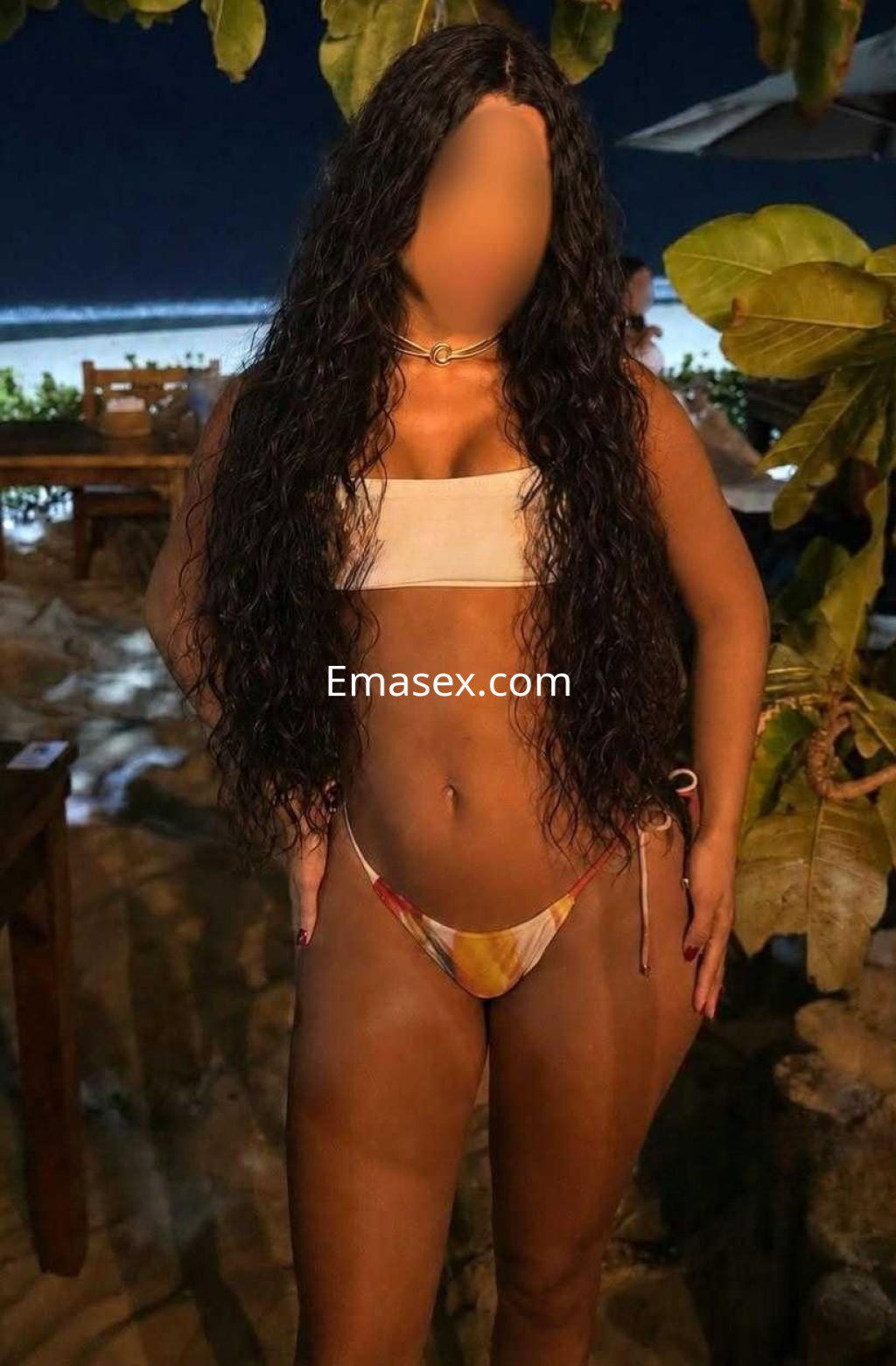 Puta en Huelva, de pie en un entorno tropical, luciendo un bikini colorido y cabello rizado y largo.
