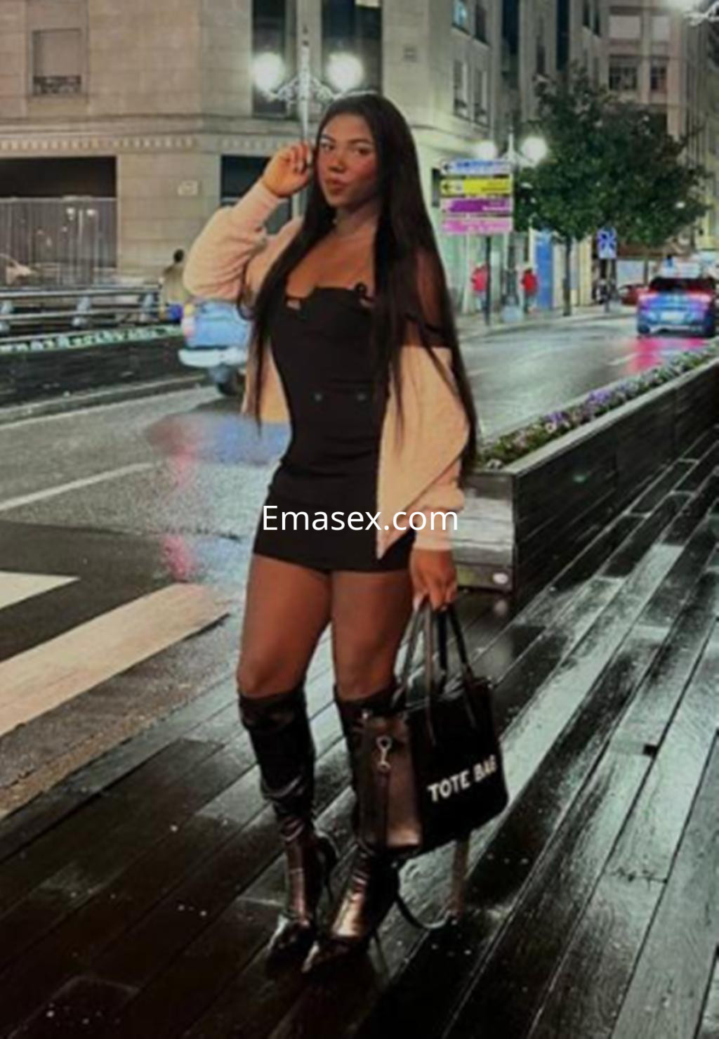 Escort trans en Logroño, vistiendo un vestido negro ajustado y botas altas, posando en la calle iluminada.