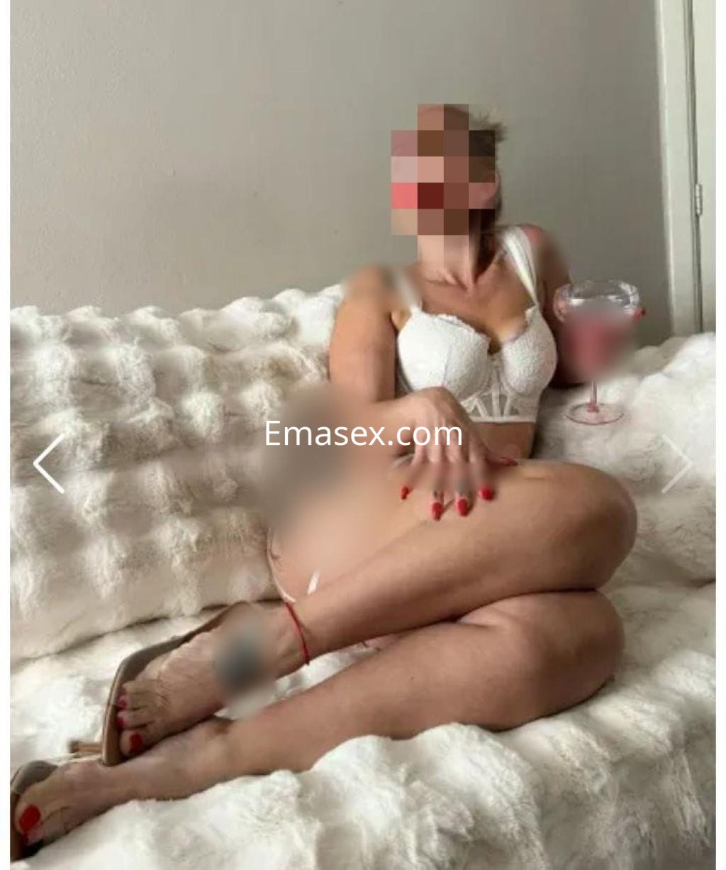 Escort en Valladolid sentada en un sofá blanco, luciendo lencería blanca y sosteniendo una bebida.
