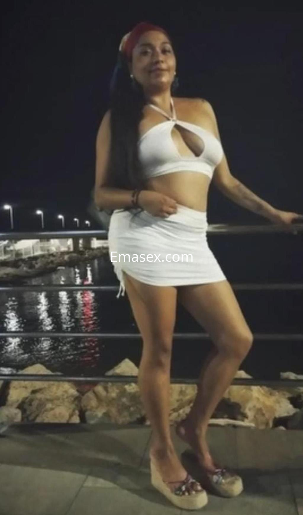La escort posa junto al mar de noche, con un vestido blanco ajustado y cabello largo, en Murcia.