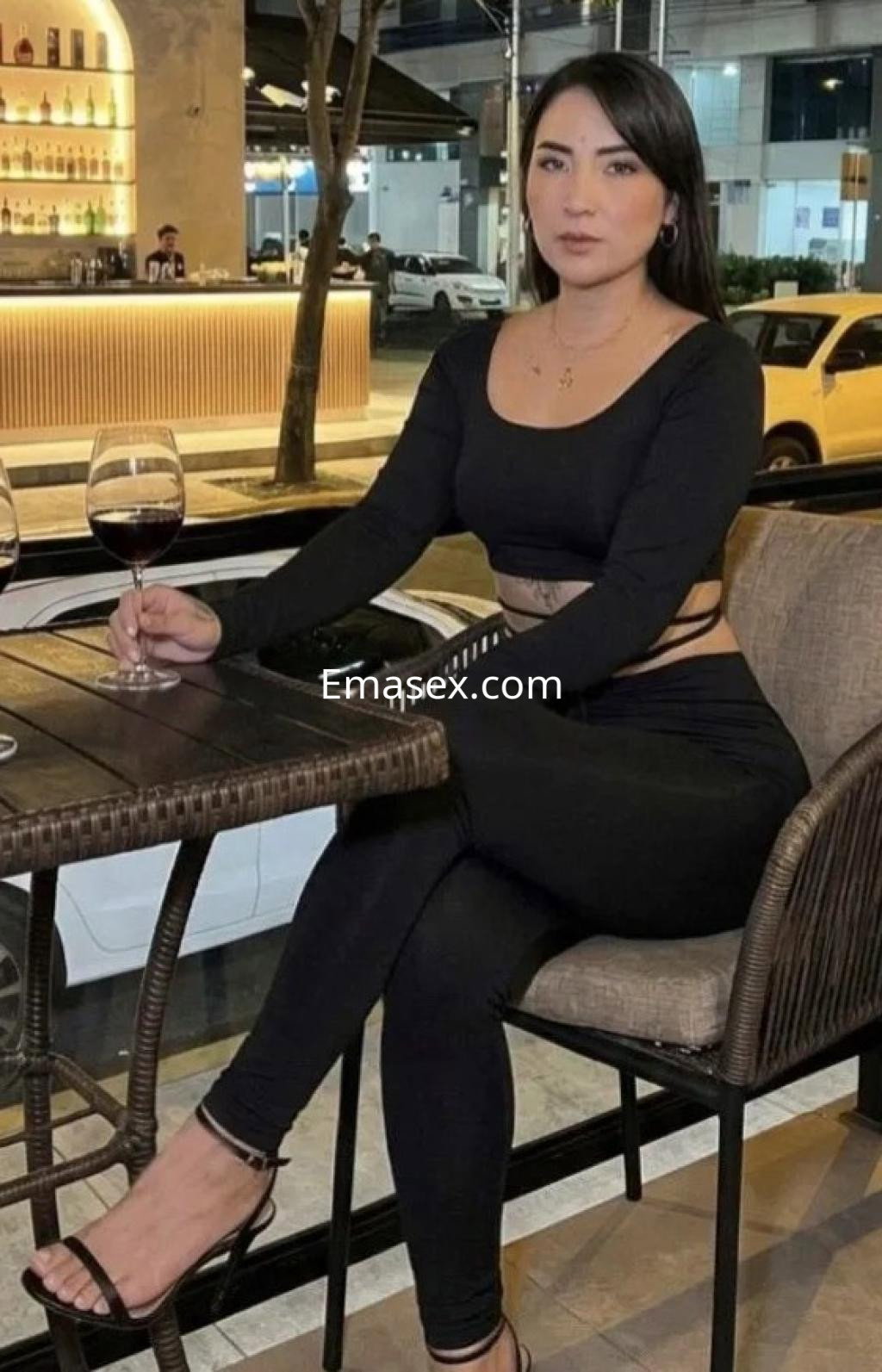 Escort en Burgos, sentada en una terraza con una copa de vino, viste un conjunto negro ajustado y tiene cabello largo.