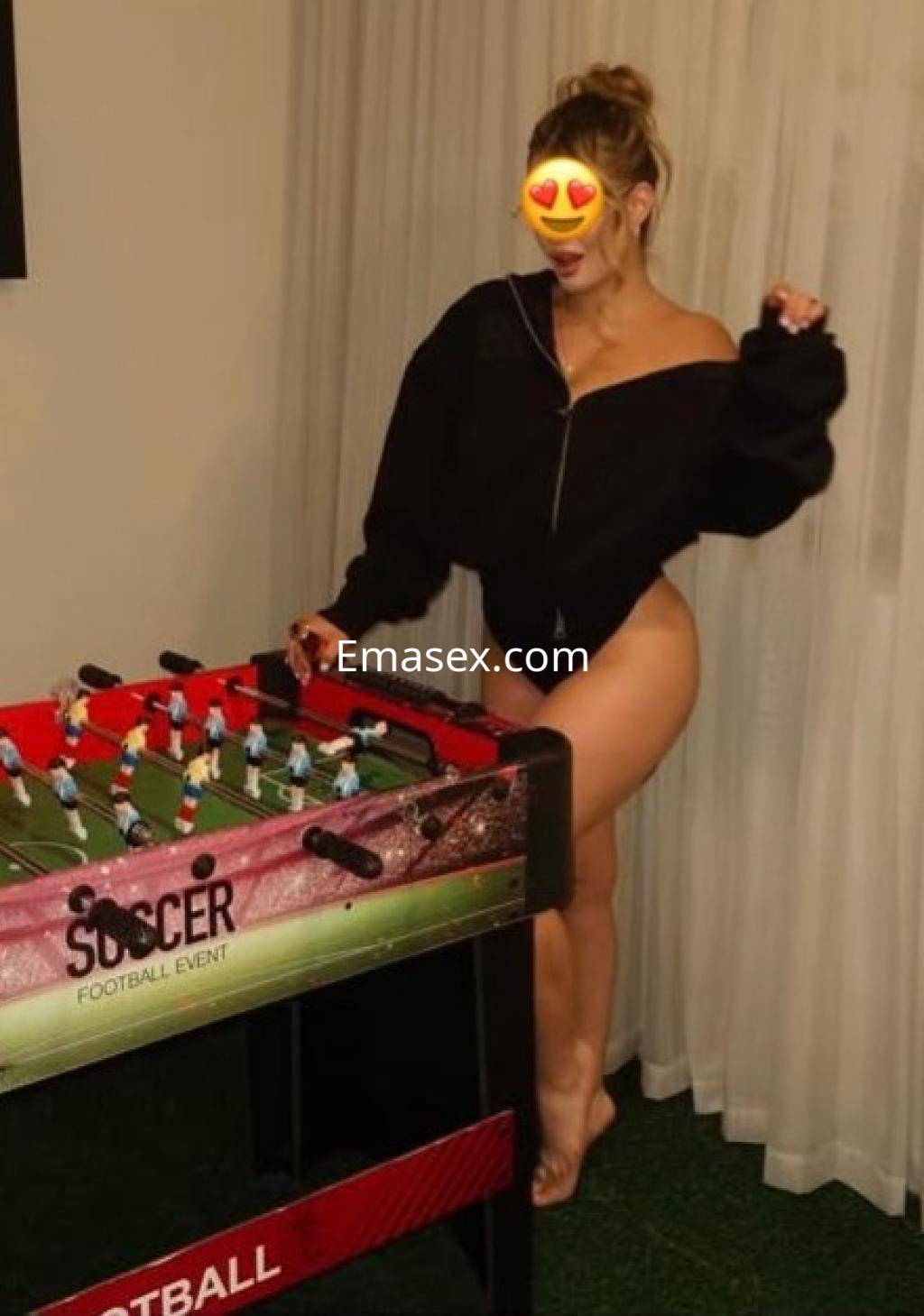 Escort en Bilbao, posando junto a un futbolín, vestida con un suéter negro y en ropa interior, luciendo una sonrisa.