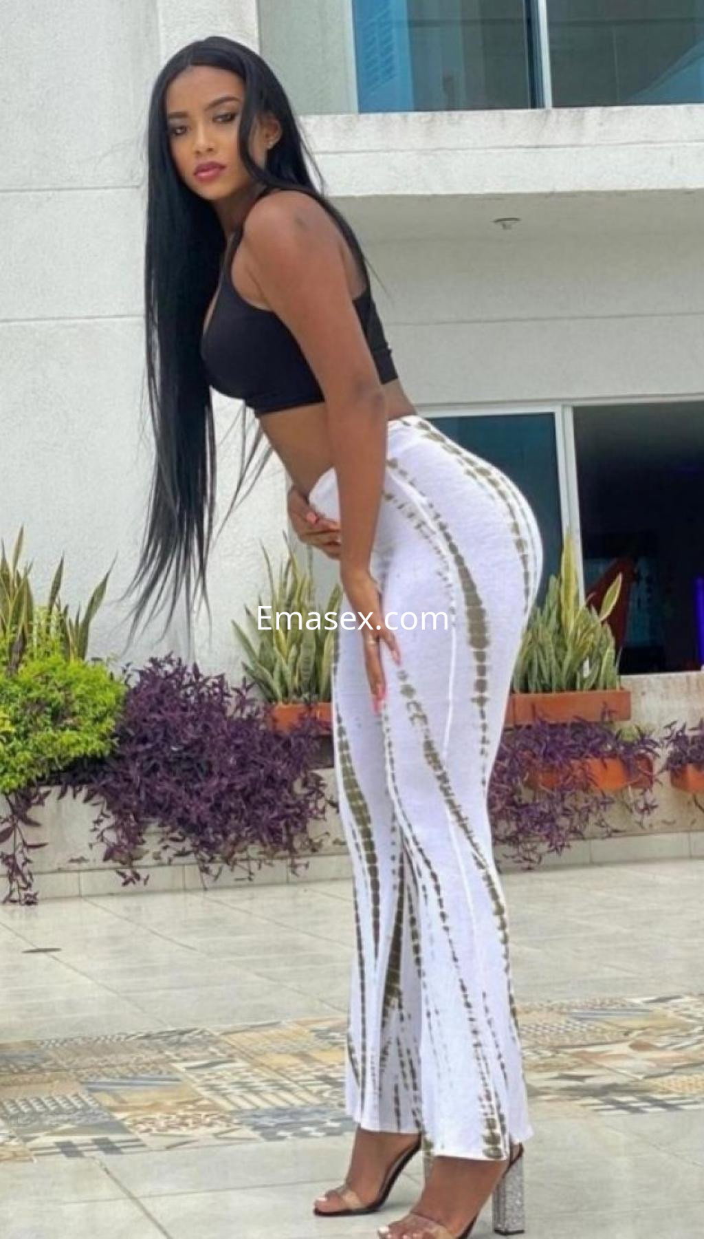 Escort en Coruña, luciendo un top negro y pantalones blancos ajustados, posando con una mano en la cadera.