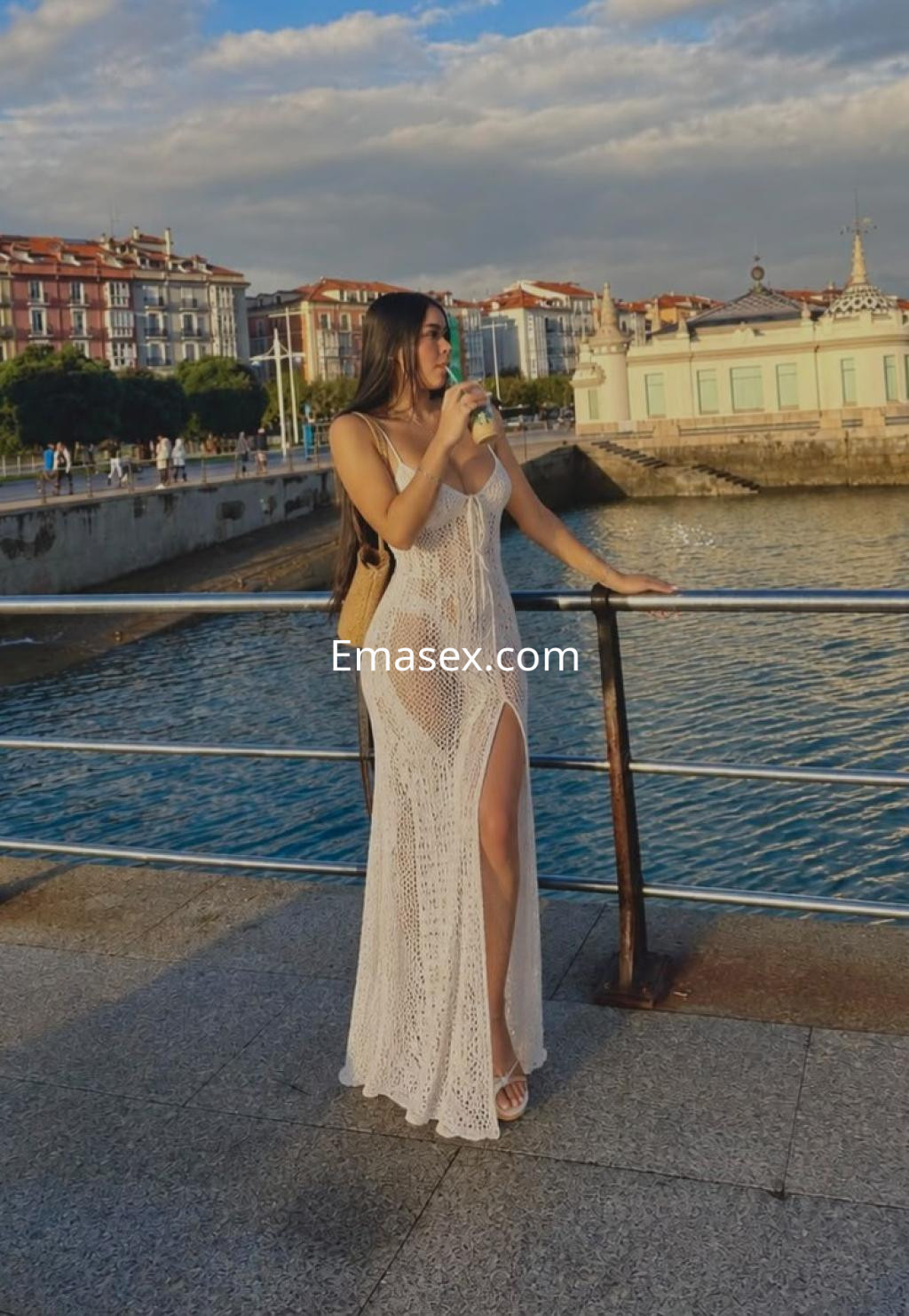 Puta en Santander, de pie junto al mar con un vestido de encaje blanco y una bebida, luciendo cabello largo.