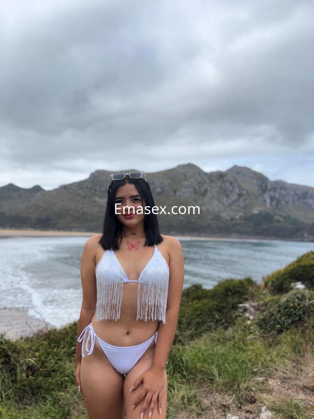 Escort en Donosti, posando junto a la playa con un bikini blanco de flecos y cabello lacio, sonriendo.