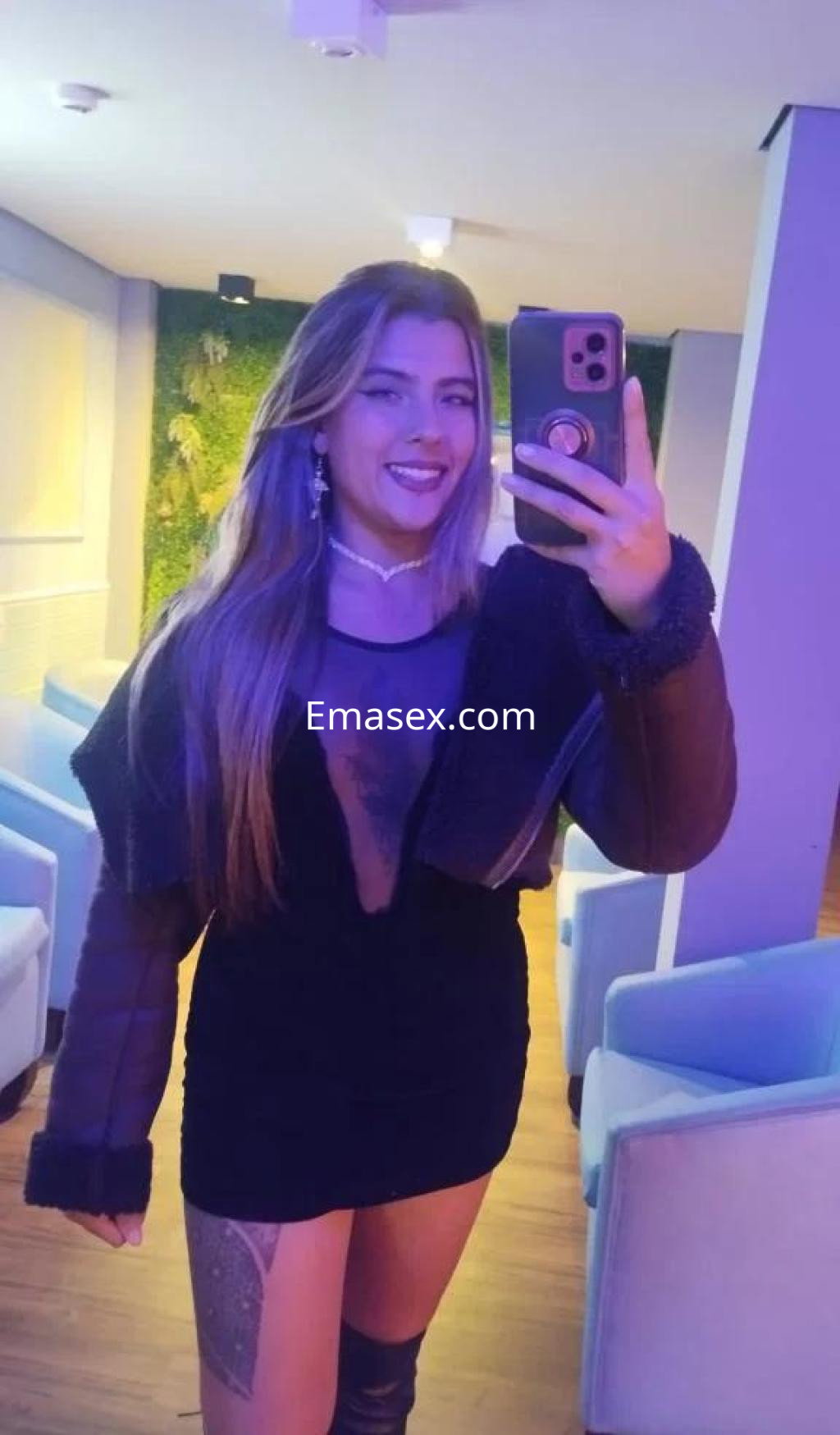 Puta posando con un vestido negro y abrigo, sonriendo mientras se toma un selfie en un entorno moderno en Girona.